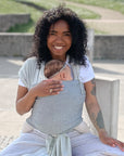 Frau trägt Baby in den Loops mit dem Übertuch "salt & pepper", sitzt auf Betonstein und Wiese im Hintergrund.