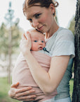 Frau trägt Baby in Loops, steht seitlich an Baum und ein Knopfgummi ist am Loop befestigt, somit kann das Baby gut herausschauen