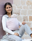 Frau trägt Baby in den Loops mit Übertuch in "blossom", sitzt vor einer Steinwand und hat ein Bein angewinkelt.