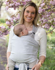 Frau trägt Baby in den Loops mit Übertuch in "moon", steht vor einem blühenden Baum und lächelt in die Kamera.