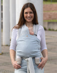 Frau trägt Baby in den Loops mit Übertuch in "sky blue", steht vor einem Betonpfeiler im Park.