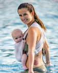 Frau trägt Baby in AQUA Loops in "sand", sie steht seitlich bis zur Hüfte im Wasser und schaut in die Kamera.