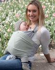 Frau trägt Baby in Loops mit Übertuch in "jade", sitzt seitlich auf einer Mauer vor einer Blumenwiese und lächelt in die Kamera.