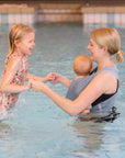Frau trägt Baby in AQUA Loops in "ocean", sie steht seitlich bis zur Hüfte im Wasser und hält die Hände von ihrem größerem Kind, das mit im Wasser ist.