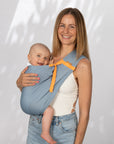 Frau trägt Baby mit einem AQUA Loop in "ocean mit orangenem Ripsband" als Hüftsitz auf der Hüfte. Beide schauen in die Kamera und sie hält eine Hand am Rücken des Babys.