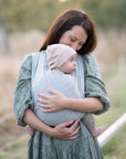 Frau trägt Baby in Loops in "greige", ohne Übertuch. Im Hintergrund Natur.