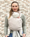 Frau trägt Baby in Loops mit Übertuch in "hazel", steht vor einer Steinmauer.