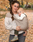 Frau trägt Kleinkind mit einem Loop als Hüfttrage auf der Hüfte und steht dabei in einem Herbstwald.