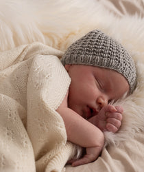 schlafendes Baby mit grauer Strickmütze, eingewickelt in einer cremefarbenen Decke. 