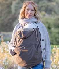 Frau mit grauem Tragecover und Schal, draußen vor Bäumen und Wiese.