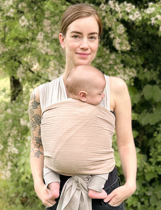 Frau trägt Baby in AQUA Loops in "sand" mit Musselin-Übertuch in "caramel", steht vor Bäumen.