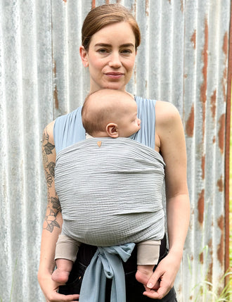 Frau trägt Baby in AQUA Loops in "ocean" mit Musselin-Übertuch in "cloud", steht vor Blechwand.